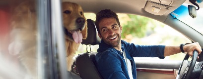 Un homme souriant conduisant une voiture avec un chien sur le siège passager.