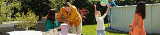 na famiglia si gode un momento insieme in un giardino soleggiato. Un uomo con una camicia gialla gioca con un bambino mentre due donne osservano e sorridono.