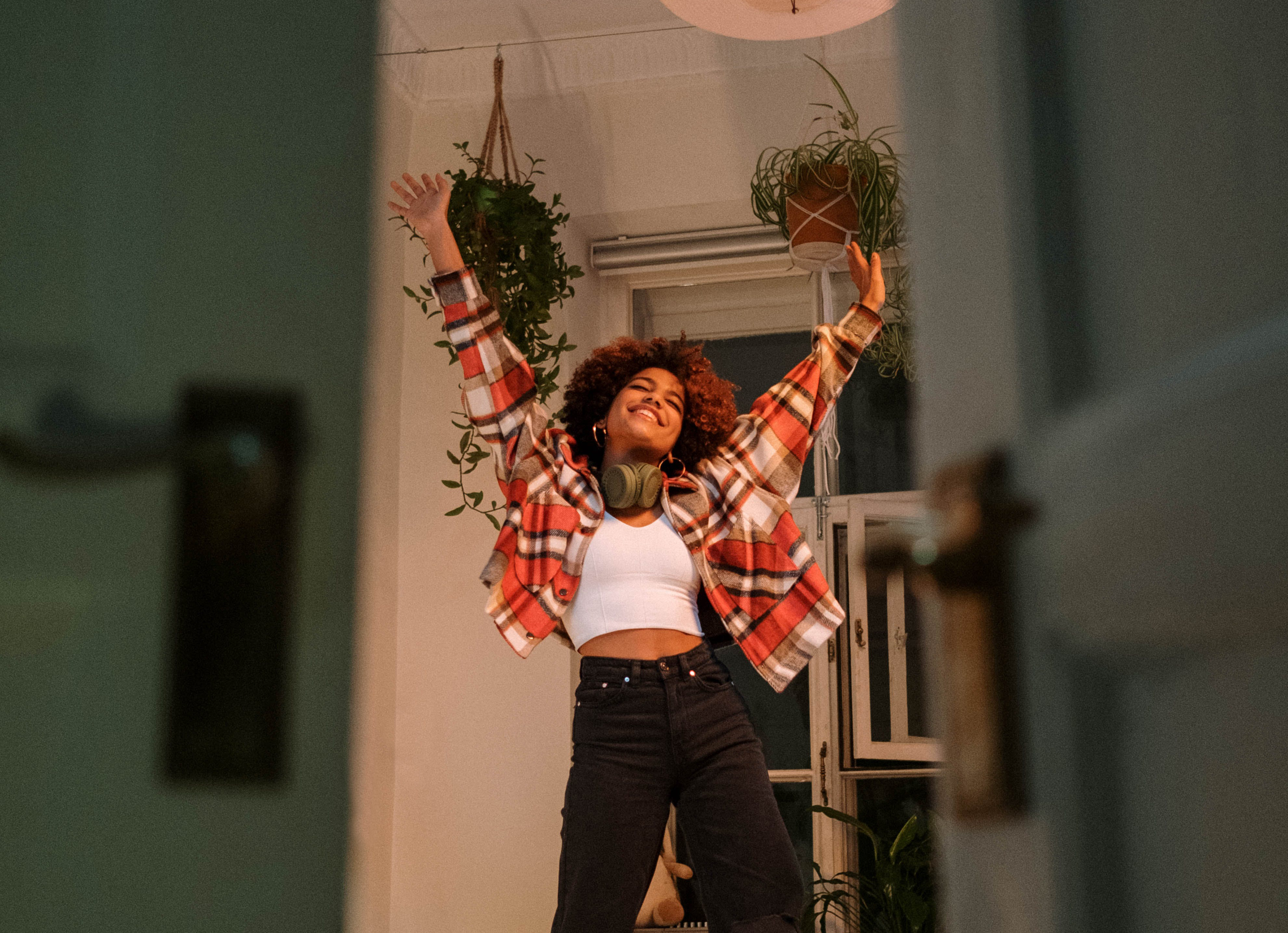 Une jeune femme joyeuse avec des cheveux afro danse chez elle, les bras levés en l'air, entourée de plantes suspendues. Elle porte un casque autour du cou et une chemise à carreaux sur un t-shirt blanc.