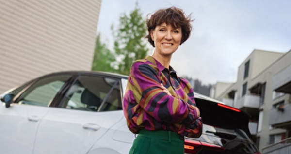 Une femme d'affaires souriante avec les bras croisés se tient devant sa voiture électrique blanche de luxe dans un environnement urbain.