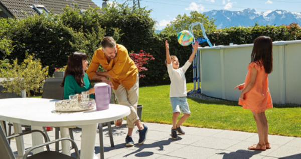 Ein Mann spielt mit Kindern in einem sonnigen Garten, mit einem Picknicktisch und Bergen im Hintergrund.