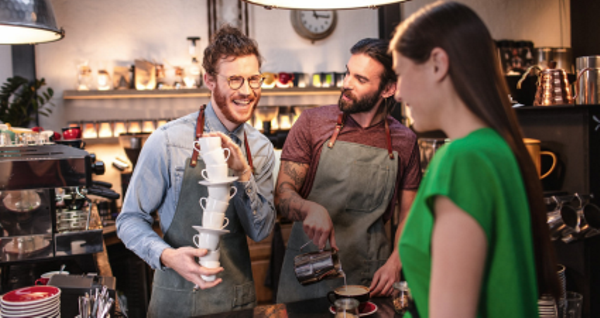 Zwei lächelnde Baristas in Schürzen interagieren mit einer Kundin in einem gemütlichen Café, einer hält einen Stapel Tassen und der andere gießt Kaffee ein.