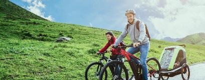 Un adulte et un enfant souriants font du vélo sur un sentier montagneux, avec une remorque à l'arrière.