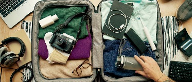 Valise ouverte avec vêtements, appareil photo, lunettes, carnet, écouteurs, brosse à dents, bouteille d’eau tenue par une main tatouée. Autour : ordi portable, carnet, écouteurs, carte.