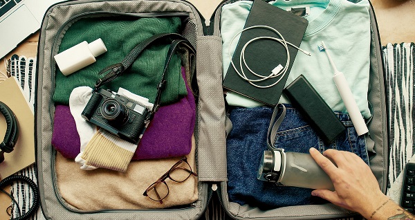 Valise ouverte avec vêtements, appareil photo, lunettes, carnet, écouteurs, brosse à dents, bouteille d’eau tenue par une main tatouée. Autour : ordi portable, carnet, écouteurs, carte.