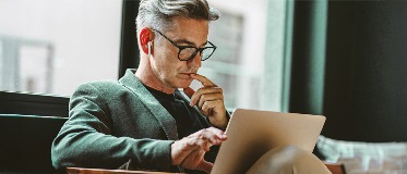Homme d’environ 40 ans assis, regardant un ordinateur portable. Il porte des lunettes, une chemise foncée et un pantalon clair.