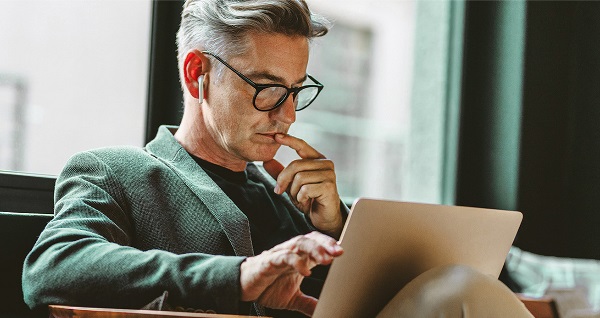 Homme d’environ 40 ans assis, regardant un ordinateur portable. Il porte des lunettes, une chemise foncée et un pantalon clair.